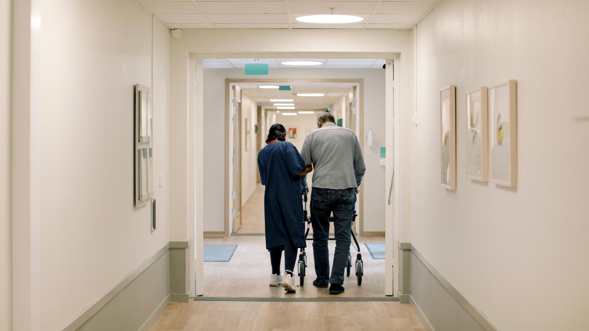 Blick in den Flur eines Pflegeheims. Von hinten sieht man eine Krankenschwester, die eine Person beim Gehen mit einer Gehhilfe unterstützt. Blick in den Flur eines Pflegeheims. Von hinten sieht man eine Krankenschwester, die eine Person beim Gehen mit einer Gehhilfe unterstützt.