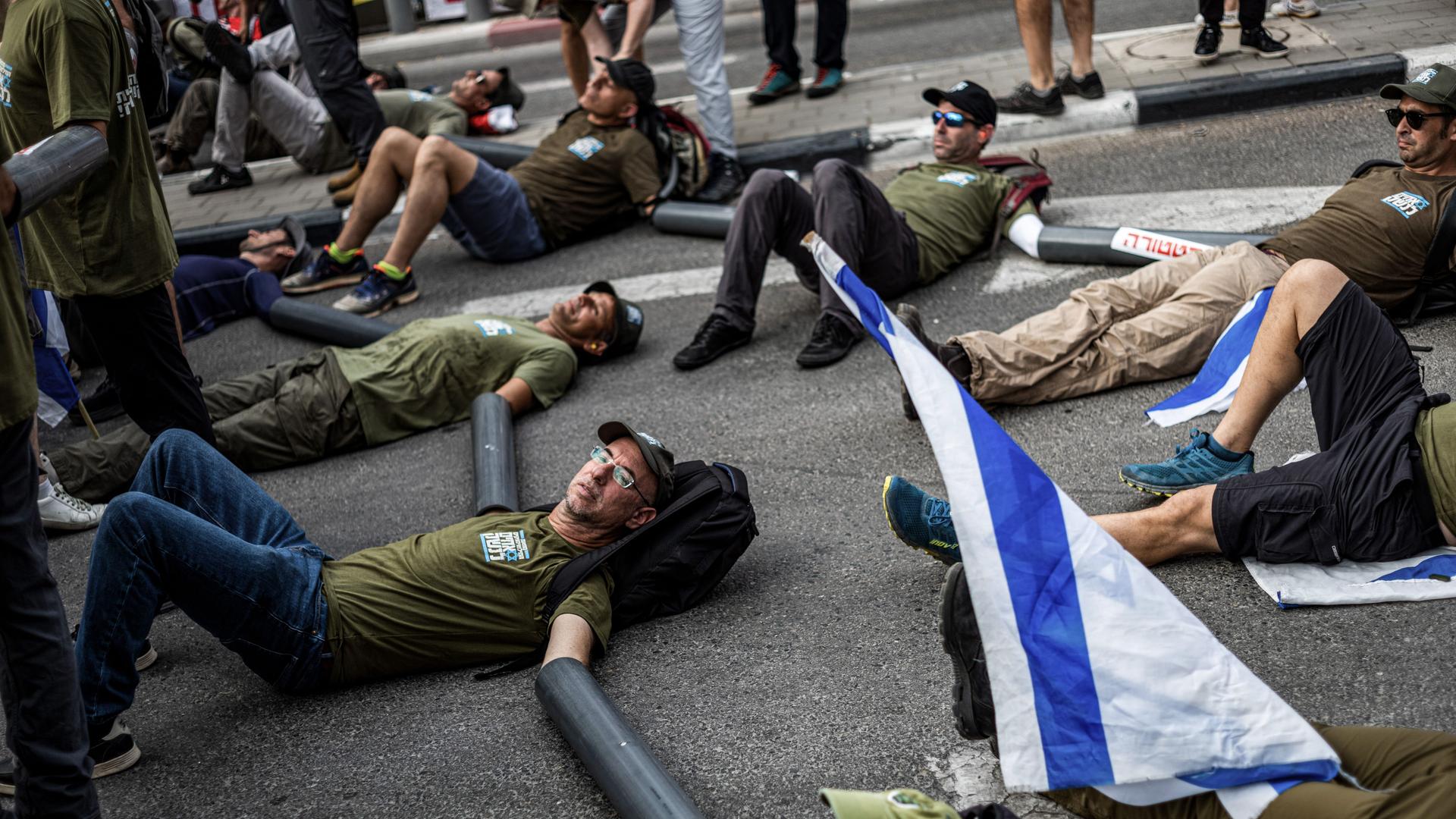 Die Militärreservisten liegen auf dem Boden einer Einfahrt, ihre Hände stecken in schwarzen Plastikschläuchen. Daneben hängt eine israelische Flagge. Die Militärreservisten liegen auf dem Boden einer Einfahrt, ihre Hände stecken in schwarzen Plastikschläuchen. Daneben hängt eine israelische Flagge.