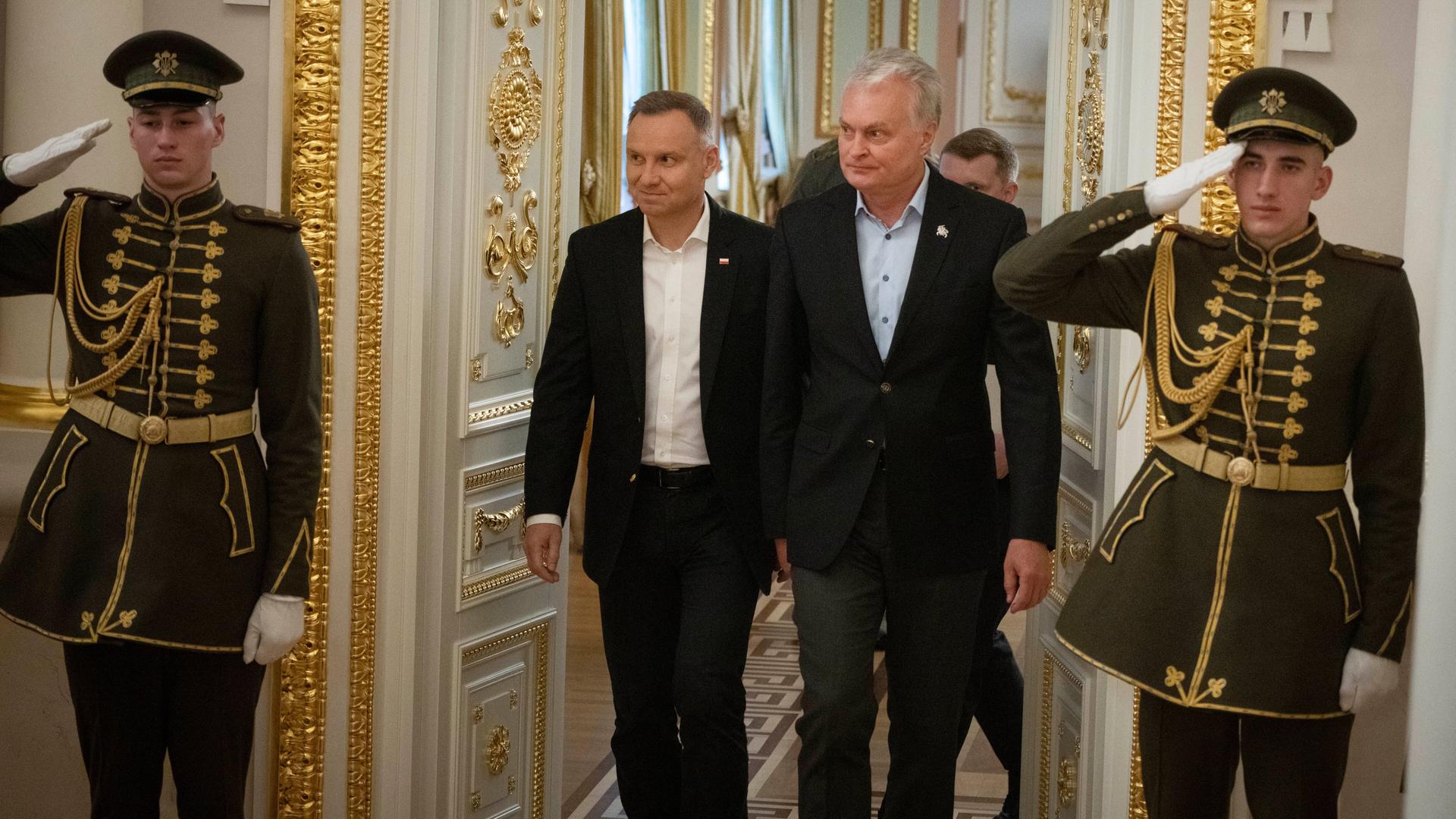 Die beiden in schwarzen Jacken treten durch eine mit Gold verzierte Tür. Rechts und links von ihnen salutiert ein Soldat in einer prächtigen dunkelgrün-goldenen Uniform. Die beiden in schwarzen Jacken treten durch eine mit Gold verzierte Tür. Rechts und links von ihnen salutiert ein Soldat in einer prächtigen dunkelgrün-goldenen Uniform.
