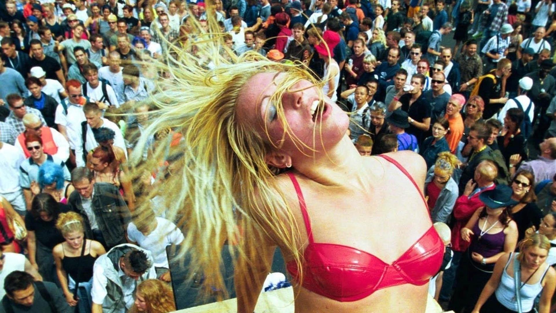 Eine blonde Frau in rosa Bikini und Plateauschuhen tanzt auf einem Loveparade-Wagen vor Hunderten Menschen. Eine blonde Frau in rosa Bikini und Plateauschuhen tanzt auf einem Loveparade-Wagen vor Hunderten Menschen.