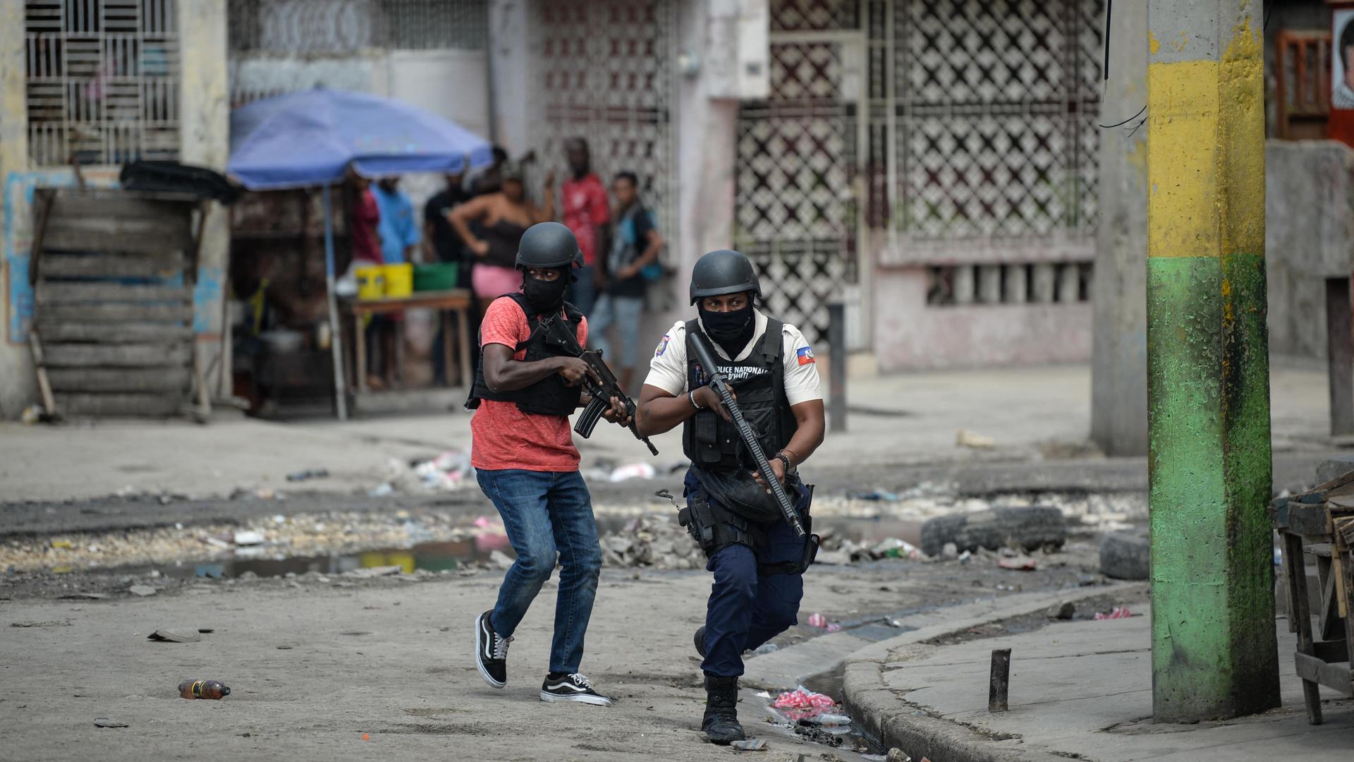 Polizisten ziehen mit im Anschlag befindlichen Waffen durch Port-au-Prince. Im Hintergrund drängen sich mehrere Menschen in einen Hauseingang. Polizisten ziehen mit im Anschlag befindlichen Waffen durch Port-au-Prince. Im Hintergrund drängen sich mehrere Menschen in einen Hauseingang.
