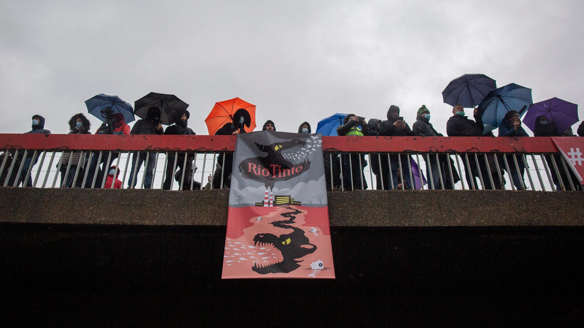Demonstranten halten auf einer Brücke ein Transparent hoch, auf dem ein illustriertes Monster zu sehen ist, das sich aus einer Fabrikhalle in die Erde frisst. Demonstranten halten auf einer Brücke ein Transparent hoch, auf dem ein illustriertes Monster zu sehen ist, das sich aus einer Fabrikhalle in die Erde frisst.