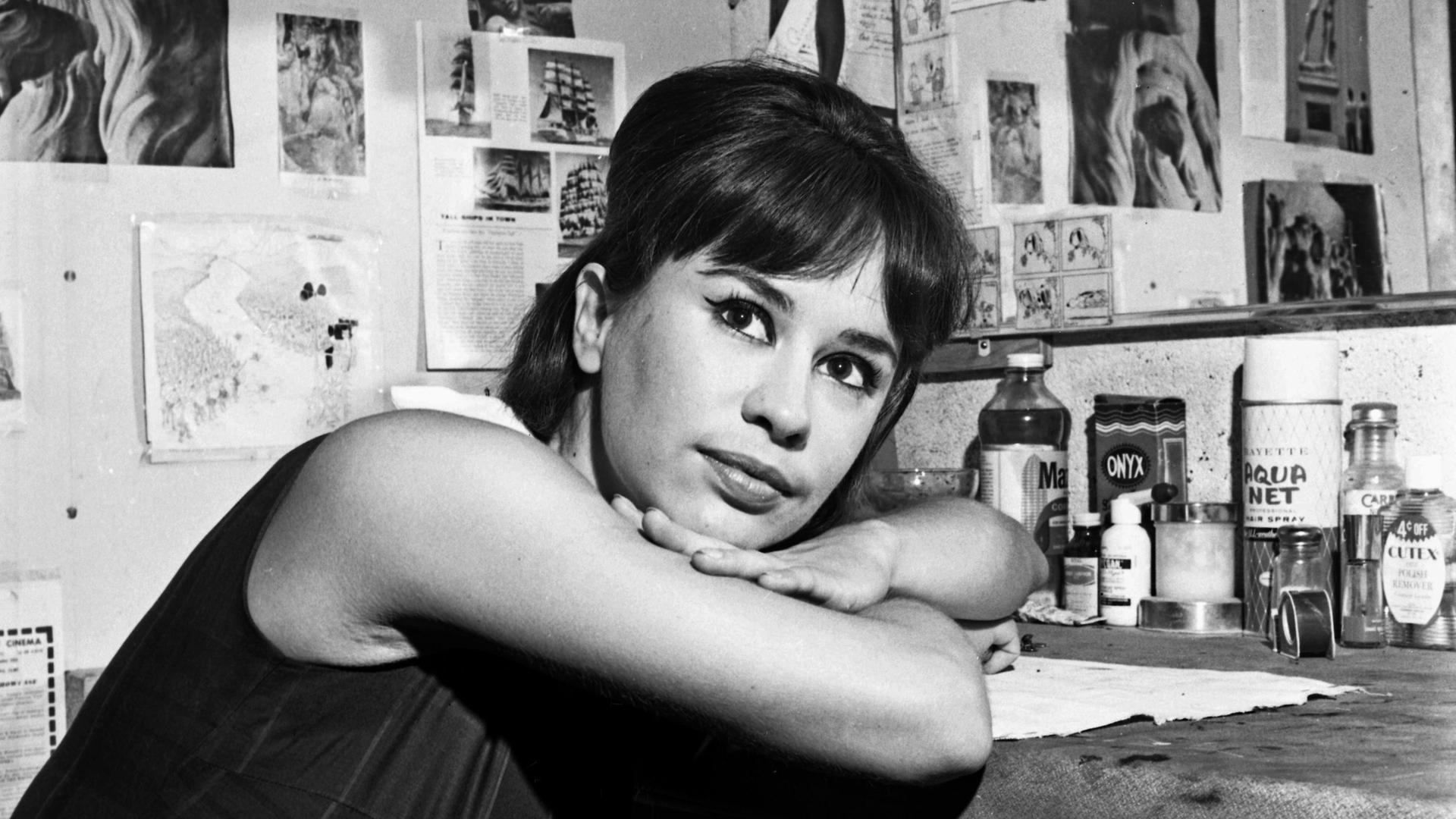 Astrud Gilberto lehnt auf einem Tisch mit vielen Gegenständen darauf und blickt auf diesem Schwarz-Weiß-Foto in die Kamera. Astrud Gilberto lehnt auf einem Tisch mit vielen Gegenständen darauf und blickt auf diesem Schwarz-Weiß-Foto in die Kamera.