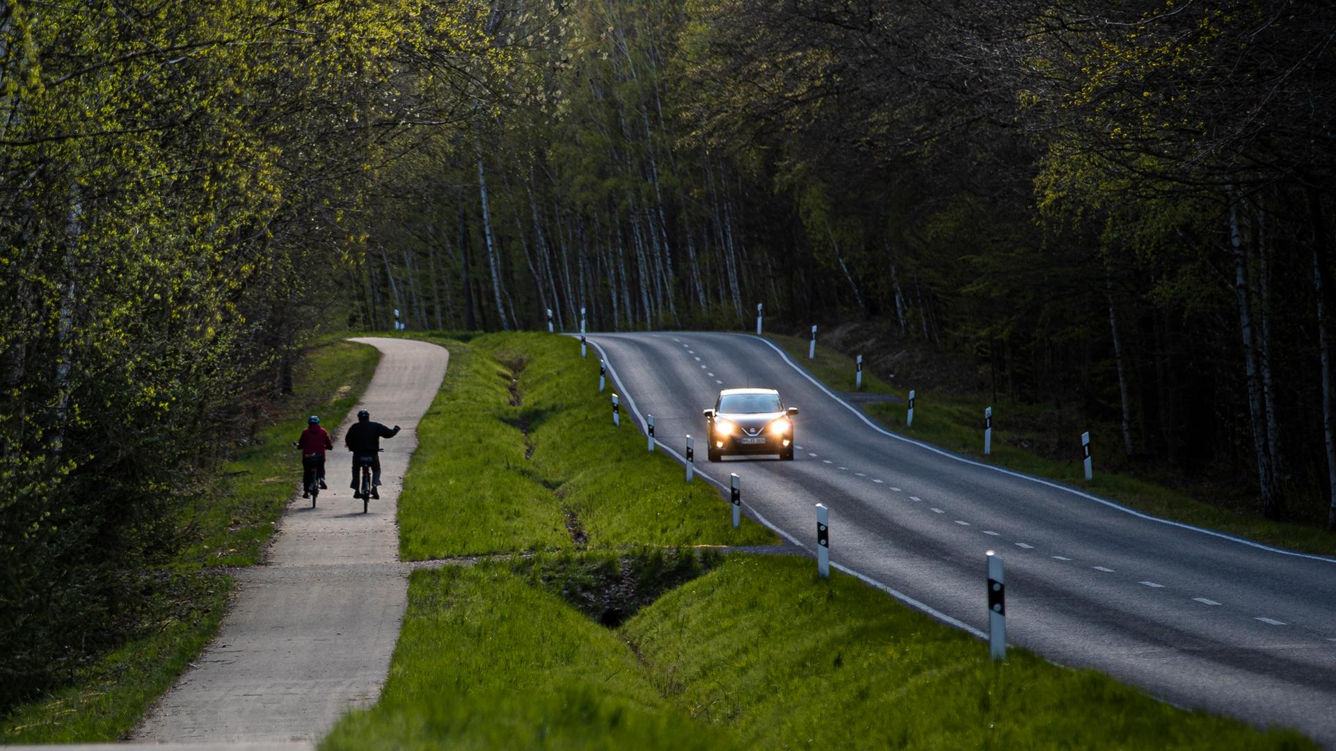 Zwei Radfahrer fahren auf einem Radweg, der direkt neben einer Landstraße verläuft. Ein Auto mit Lichtern fährt auf der Landstraße. Der rechte Radfahrer salutiert in Richtung des Autos. Zwei Radfahrer fahren auf einem Radweg, der direkt neben einer Landstraße verläuft. Ein Auto mit Lichtern fährt auf der Landstraße. Der rechte Radfahrer salutiert in Richtung des Autos.