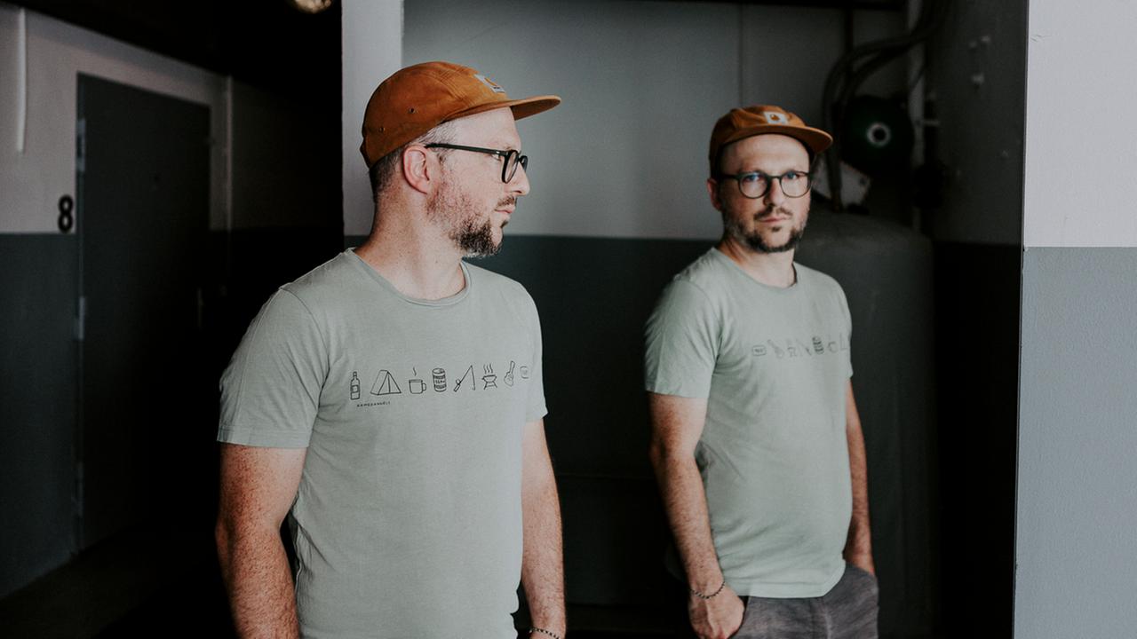 Ein Mann in T-Shirt mit Brille und , orangenem Käppi steht in einem Heizungskeller und schaut so seitlich in einen Spiegel, dass sein Gesicht den Betrachter aus dem Spiegel anblickt. Ein Mann in T-Shirt mit Brille und , orangenem Käppi steht in einem Heizungskeller und schaut so seitlich in einen Spiegel, dass sein Gesicht den Betrachter aus dem Spiegel anblickt.