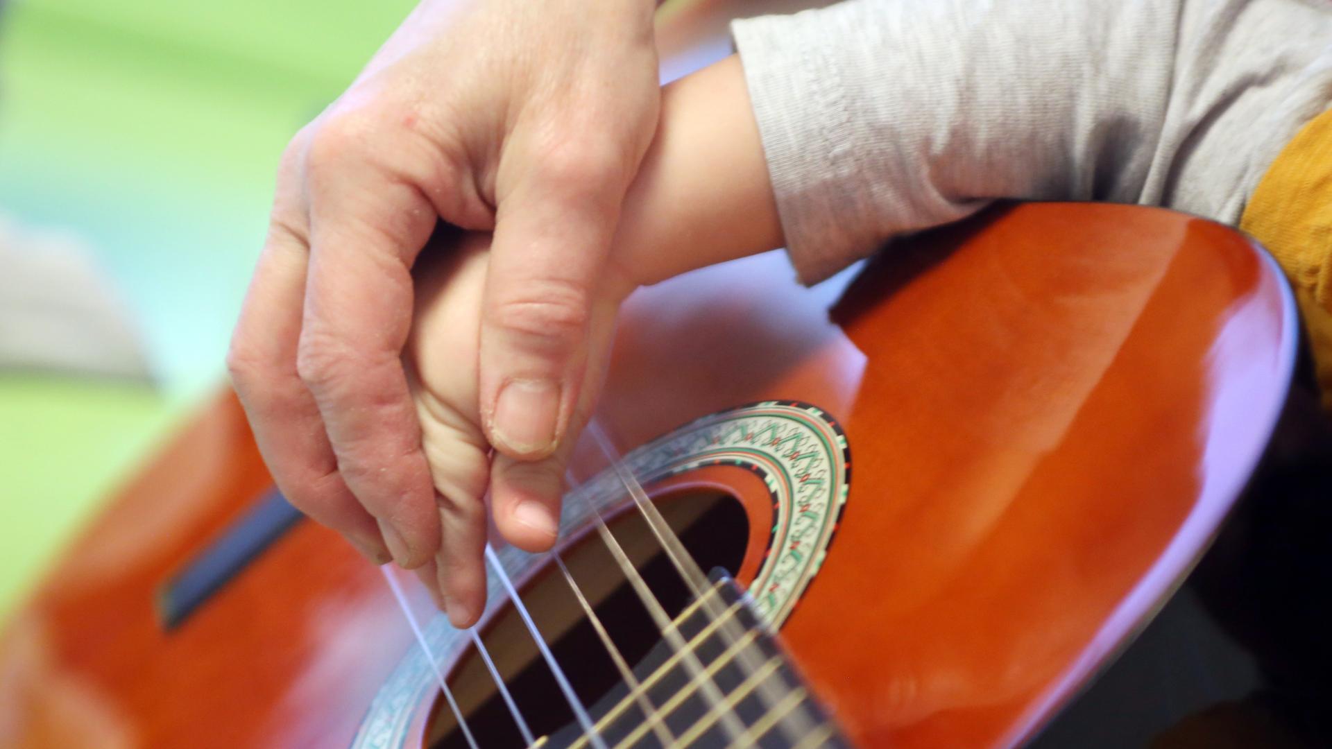Eine Erwachsenhand hilft einer Kinderhand beim Gitarrenspiel. 