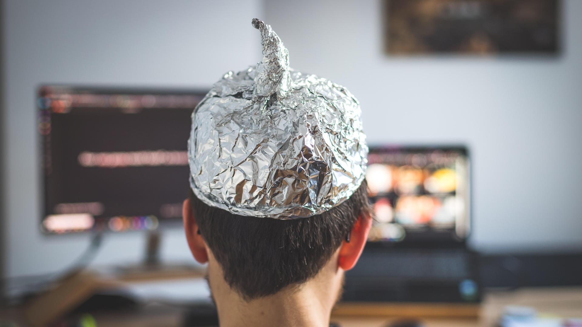 Ein Mann sitzt vor zwei Bildschirmen und trägt eine Aluminiumkappe. Auf den Bildschirmen sind verschwommene Inhalte zu erkennen. Ein Mann sitzt vor zwei Bildschirmen und trägt eine Aluminiumkappe. Auf den Bildschirmen sind verschwommene Inhalte zu erkennen.