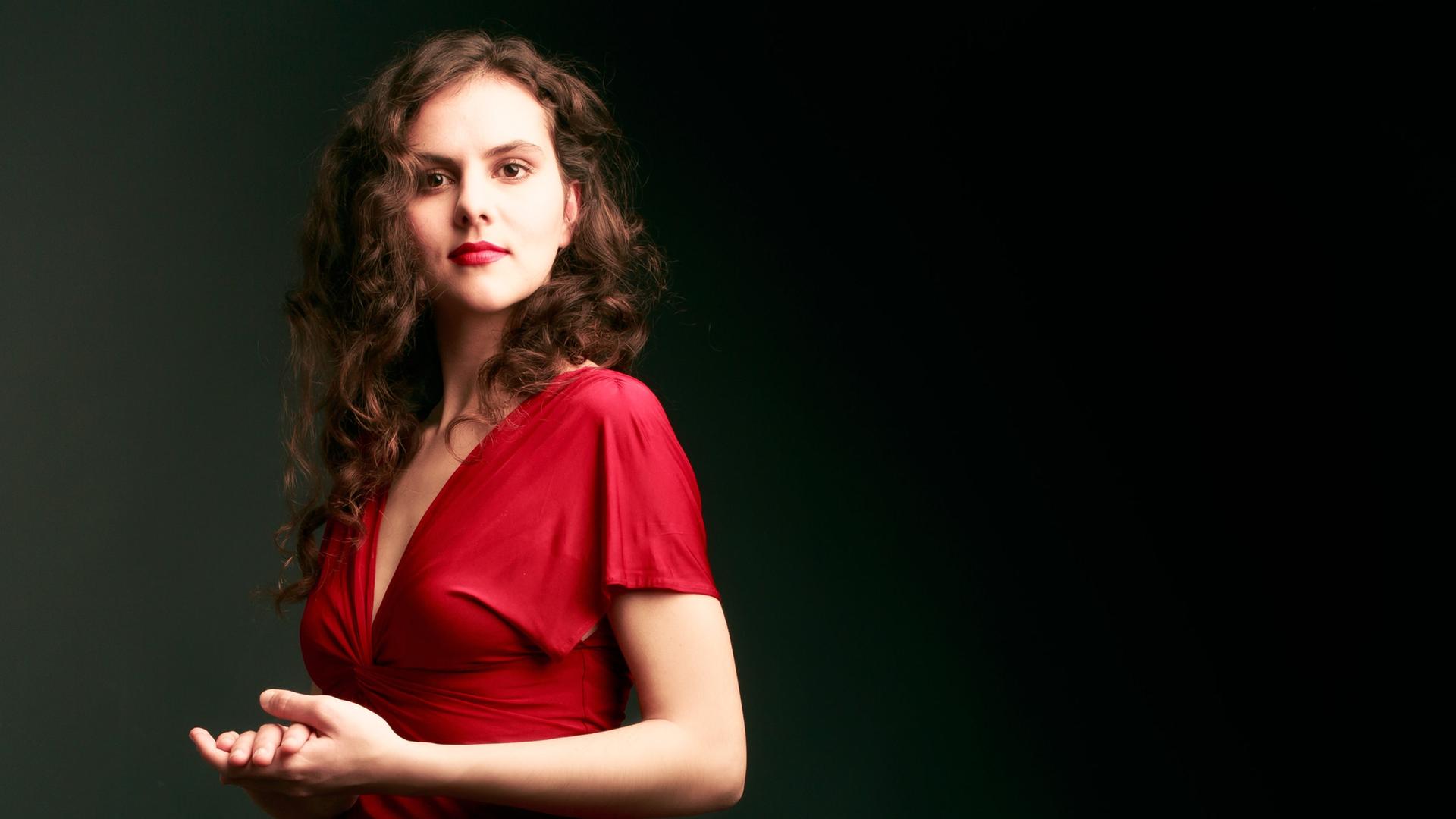 Eine Frau steht seitlich zur Kamera. die Hände vor dem Körper ineinander gelegt. Sie trägt ein rotes Kleid und hat dunkle lange Locken. Eine Frau steht seitlich zur Kamera. die Hände vor dem Körper ineinander gelegt. Sie trägt ein rotes Kleid und hat dunkle lange Locken.