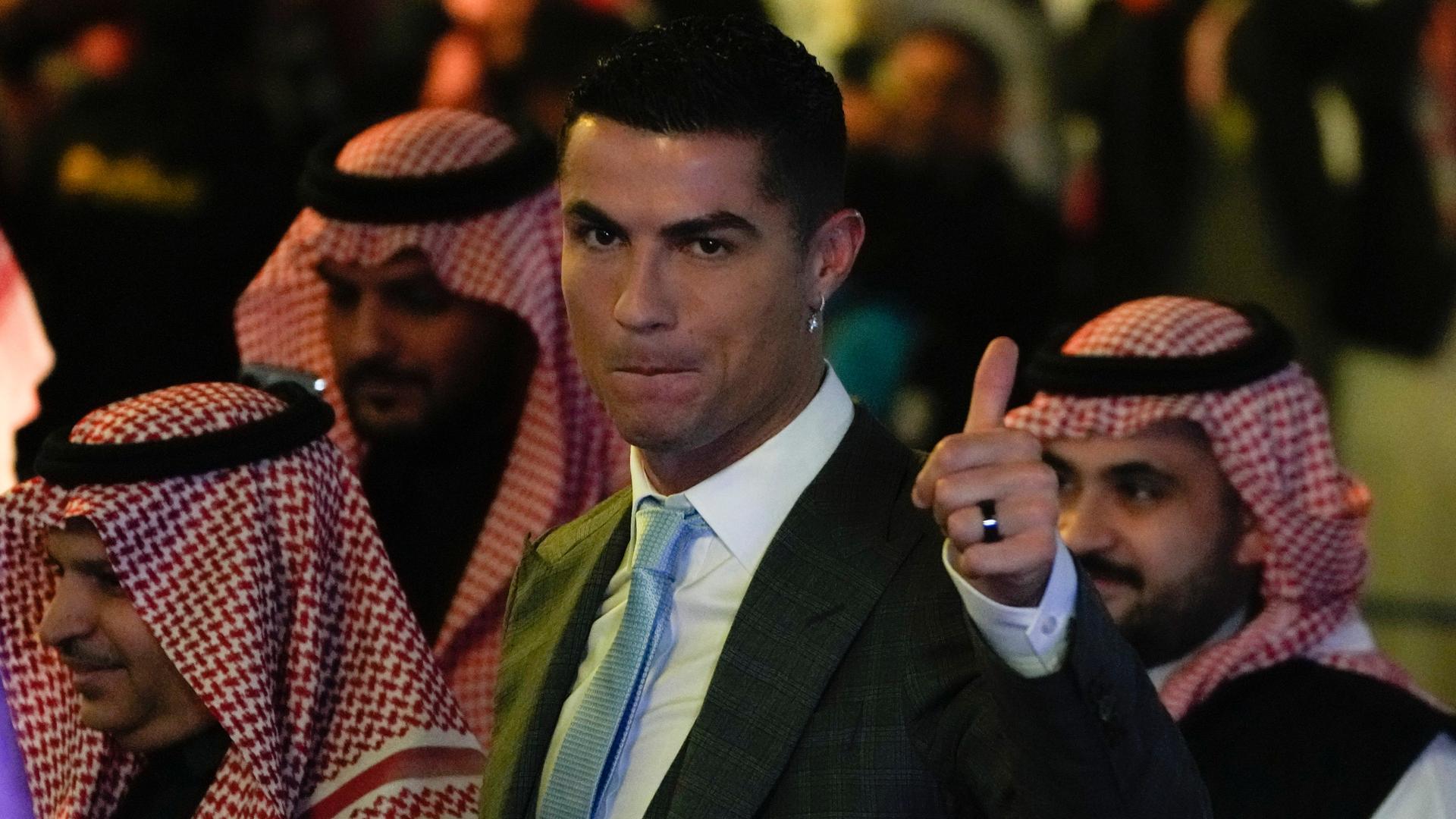 Das Foto zeigt den Fußball-Spieler Cristiano Ronaldo im Anzug. Im Hinter-Grund sieht man Männer aus dem Land Saudi-Arabien mit typischem Kopf-Tuch.