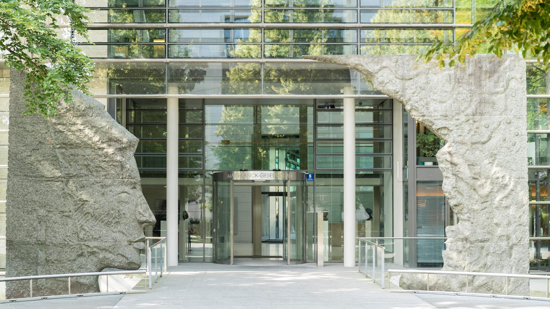 Blick auf den Eingang der Max-Planck-Gesellschaft in München, einem Glasgebäude welches am Eingang von zwei Skulpturen umrahmt wird Blick auf den Eingang der Max-Planck-Gesellschaft in München, einem Glasgebäude welches am Eingang von zwei Skulpturen umrahmt wird