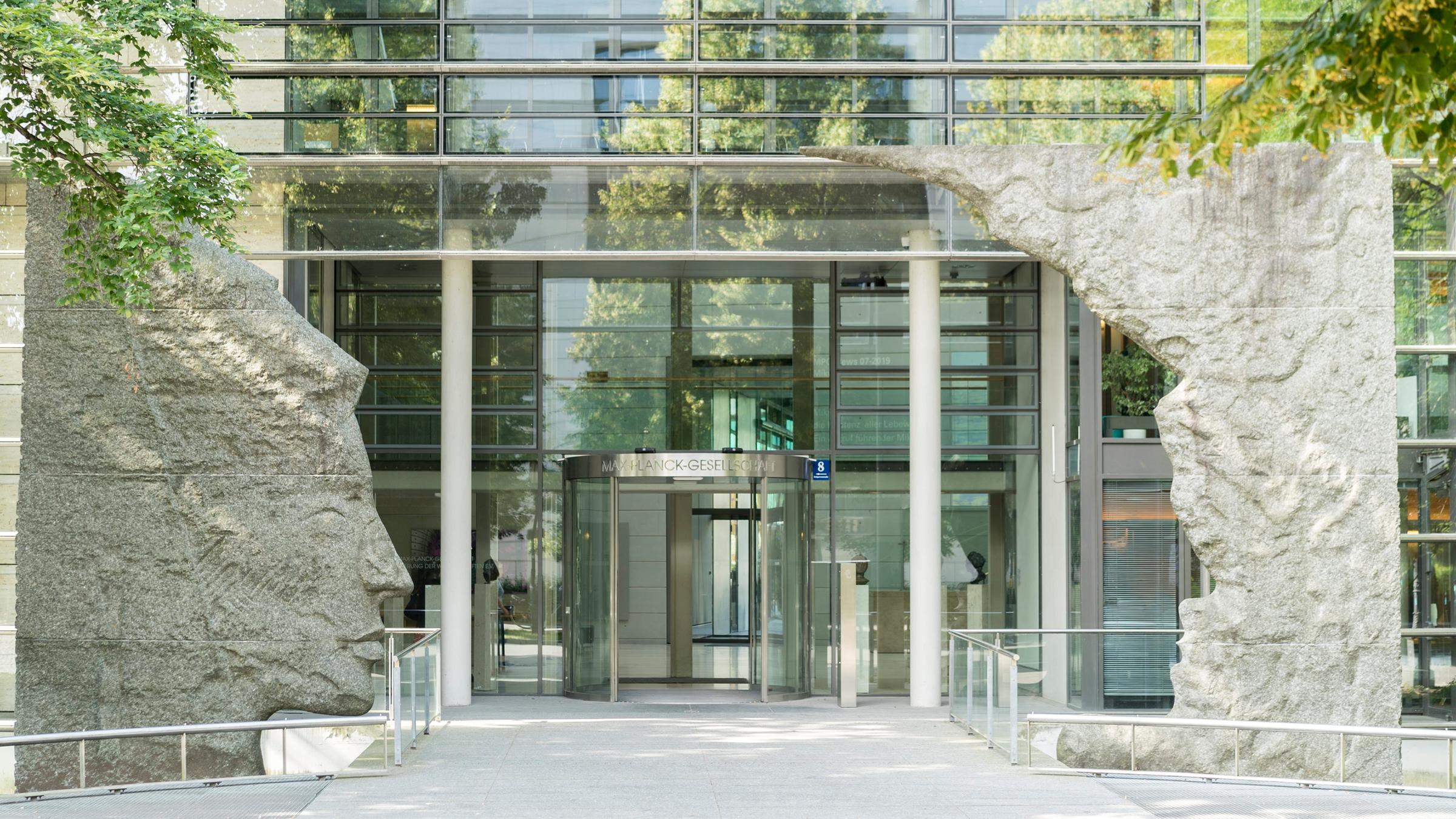 Blick auf den Eingang der Max-Planck-Gesellschaft in München, einem Glasgebäude welches am Eingang von zwei Skulpturen umrahmt wird Blick auf den Eingang der Max-Planck-Gesellschaft in München, einem Glasgebäude welches am Eingang von zwei Skulpturen umrahmt wird