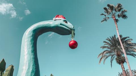 Eine großer Dinosaurie mit Weihnachtsmütze und einer roten Baumkugel im Mund überragt Palmen und andere Bäume.
