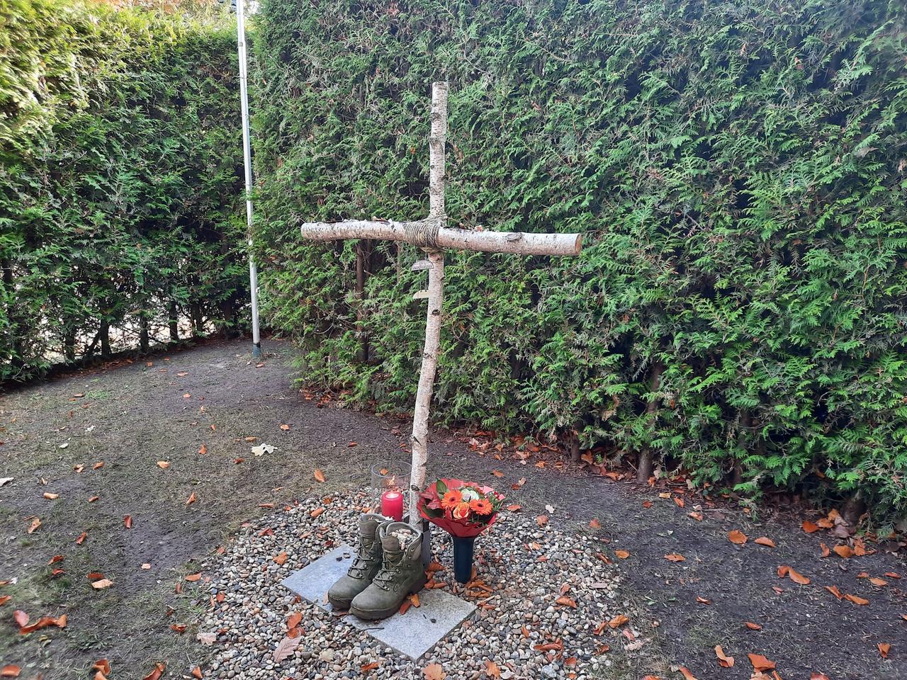 Vor einem Holzkreuz steht ein Paar robuste Stiefel, befüllt mit Kieselsteinen. Daneben steht eine Kerze und ein Vase mit Blumen. Vor einem Holzkreuz steht ein Paar robuste Stiefel, befüllt mit Kieselsteinen. Daneben steht eine Kerze und ein Vase mit Blumen.