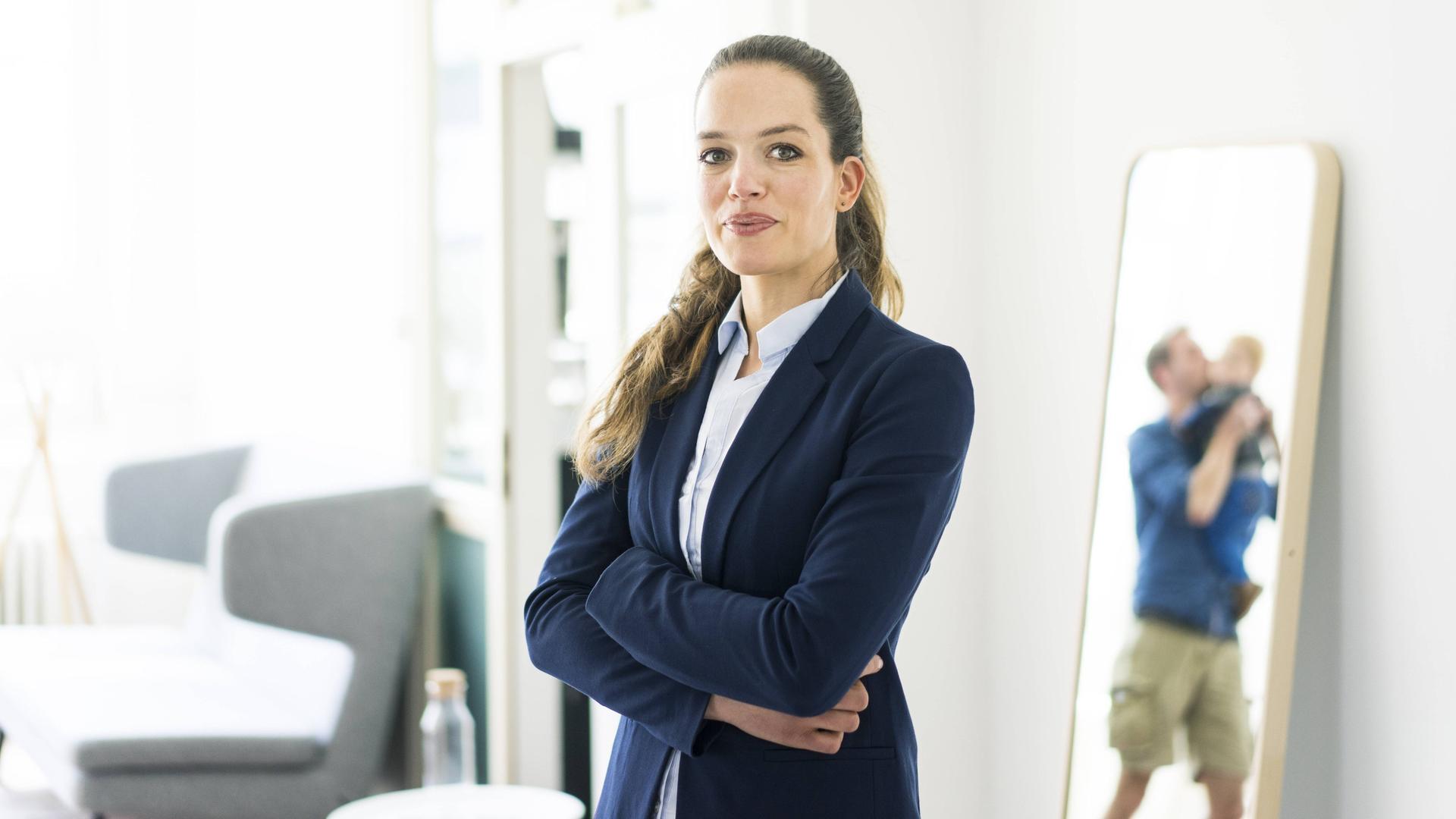 Eine Frau mit Business-Outfit steht neben einem Spiegel, in dem ein Mann mit Kleinkind zu sehen ist. Eine Frau mit Business-Outfit steht neben einem Spiegel, in dem ein Mann mit Kleinkind zu sehen ist.