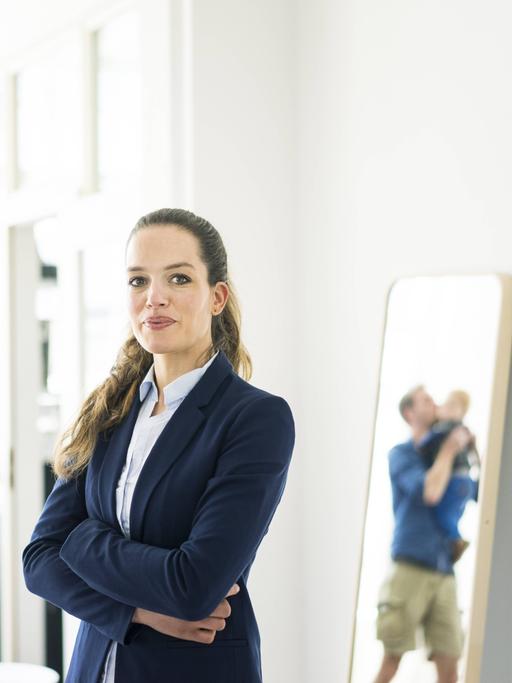 Eine Frau mit Business-Outfit steht neben einem Spiegel, in dem ein Mann mit Kleinkind zu sehen ist.