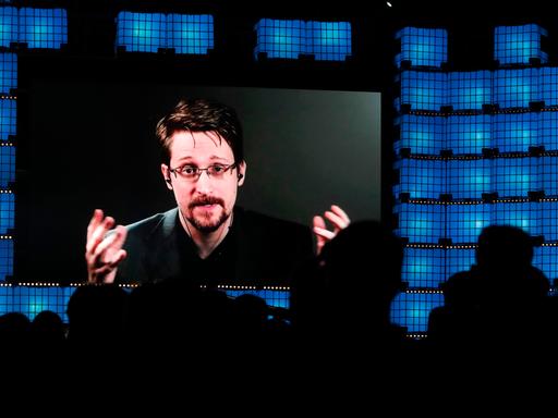 Ein Mann mit Bart und Brille ist auf einem großen Bildschirm zu sehen. Er spricht zu einem Publikum, das als Schattenriss im Vordergrund zu erkennen ist.