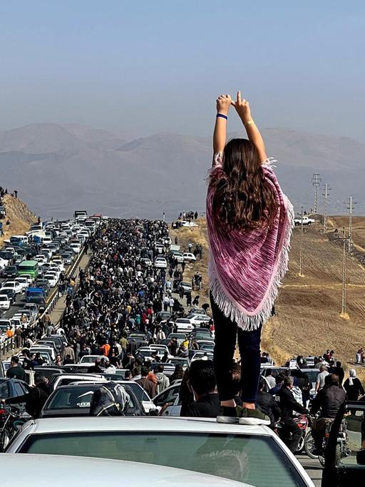 Das Foto zeigt einen Demonstrationszug im Iran, im Vordergrund eine Frau ohne Kopftuch von hinten.