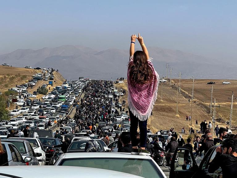 Das Foto zeigt einen Demonstrationszug im Iran, im Vordergrund eine Frau ohne Kopftuch von hinten.