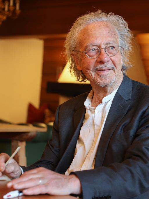 Autor Peter Handke sitzt bei einem Kartenspiel an einem Tisch und hält einen Stift in der Hand.