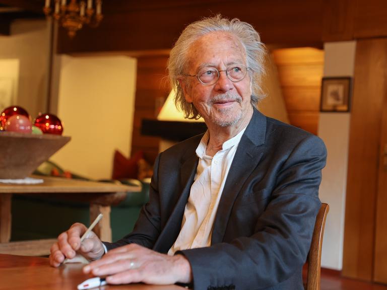 Autor Peter Handke sitzt bei einem Kartenspiel an einem Tisch und hält einen Stift in der Hand.