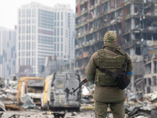 Ein ukrainischer Soldat steht mit einem Waffe in der Hand vor einem zerstörten Gebäude in Kiew. Er dreht dem Betrachter den Rücken zu.