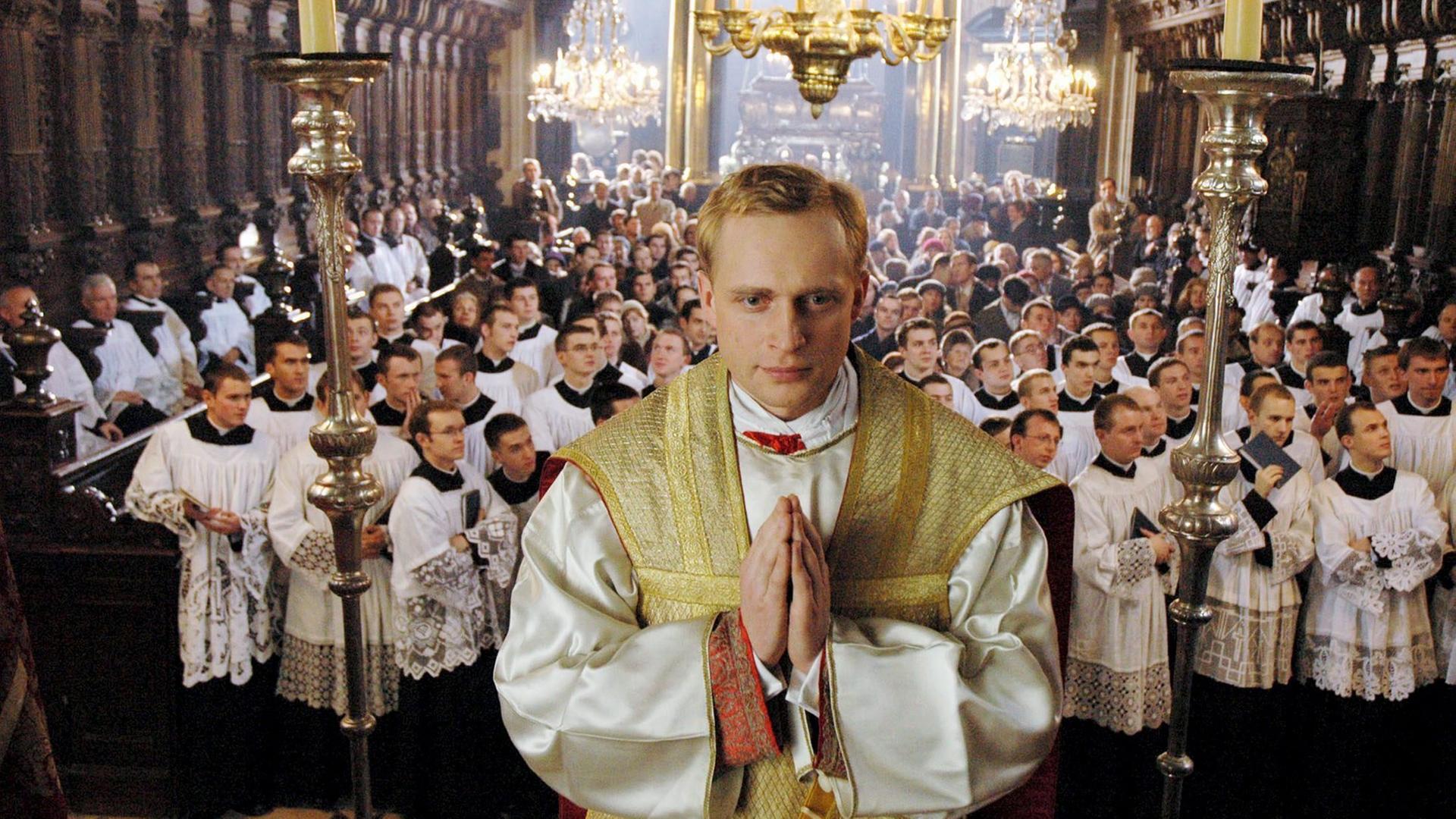  Piotr Adamczyk als Karol Wojtyla steht erhöht mit zum Gebet gefalteten Händen in einer Kirche, hinter ihm schauen viele weitere Männer mit weißen Gewändern in eine Richtung.