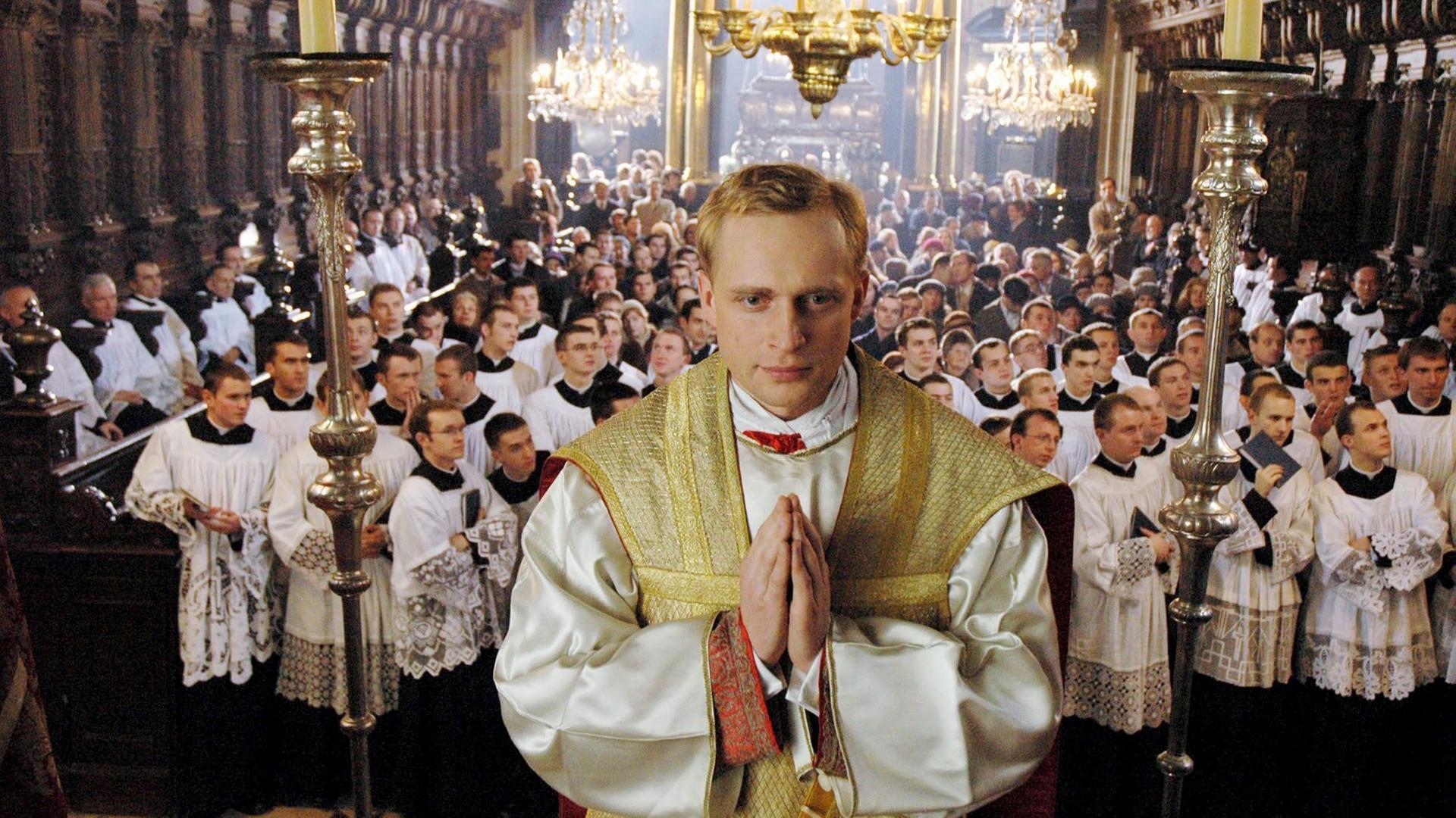 Piotr Adamczyk als Karol Wojtyla steht erhöht mit zum Gebet gefalteten Händen in einer Kirche, hinter ihm schauen viele weitere Männer mit weißen Gewändern in eine Richtung. Piotr Adamczyk als Karol Wojtyla steht erhöht mit zum Gebet gefalteten Händen in einer Kirche, hinter ihm schauen viele weitere Männer mit weißen Gewändern in eine Richtung.