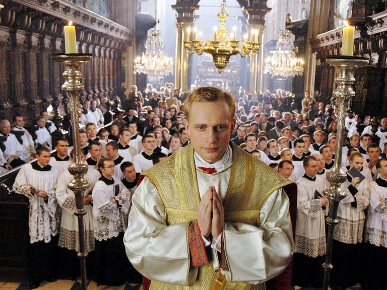  Piotr Adamczyk als Karol Wojtyla steht erhöht mit zum Gebet gefalteten Händen in einer Kirche, hinter ihm schauen viele weitere Männer mit weißen Gewändern in eine Richtung.