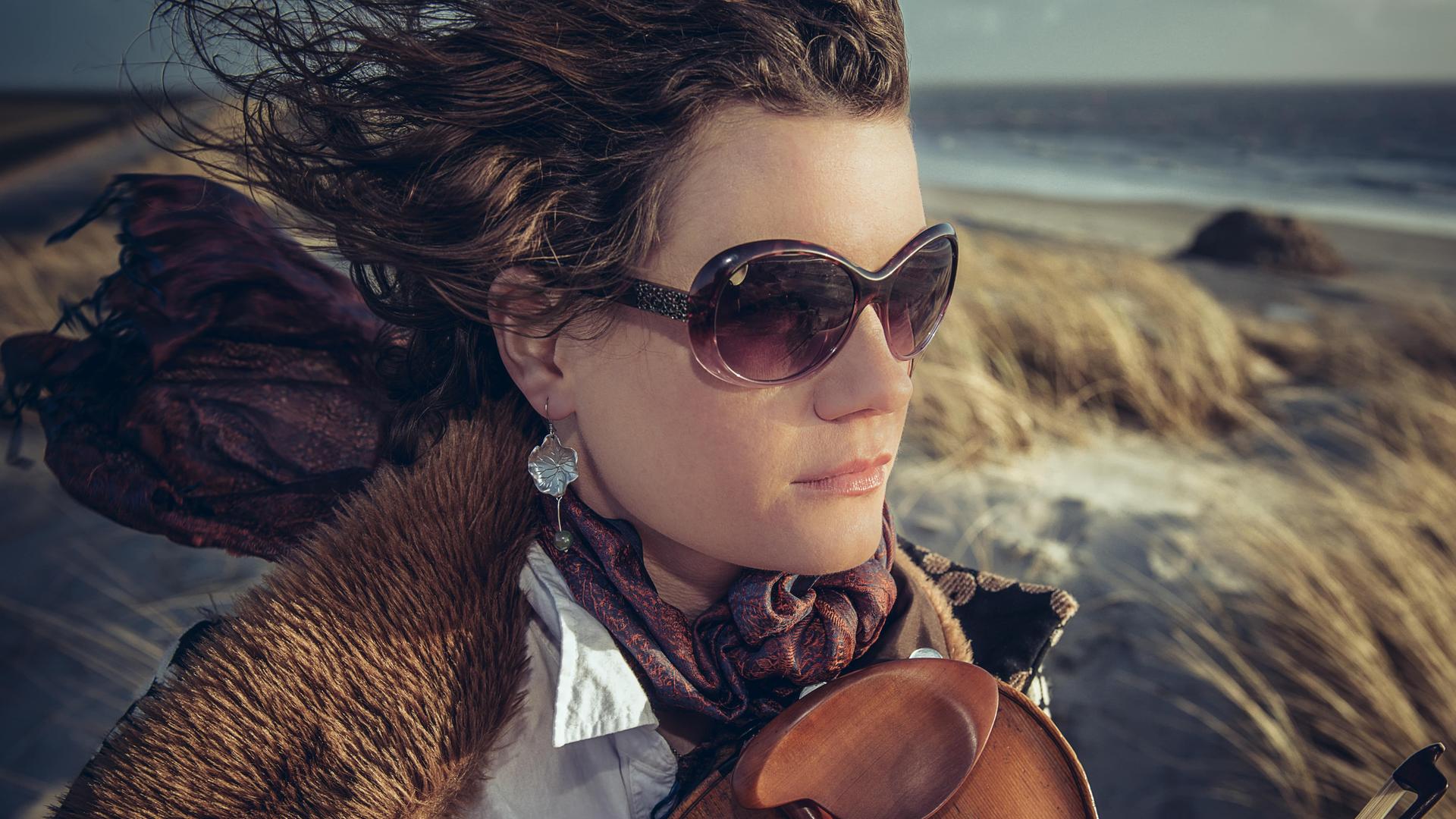 Eine Frau trägt Sonnenbrille und eine Jacke mit Pelzkragen über einer weißen Bluse. Der Wind weht ihre Haare und den buten Schal nach hinten. Auf ihrer Brust liegt locker eine Geige auf. Im Hintergrund sind Dünen und ein Meeresstrand zu sehen. Eine Frau trägt Sonnenbrille und eine Jacke mit Pelzkragen über einer weißen Bluse. Der Wind weht ihre Haare und den buten Schal nach hinten. Auf ihrer Brust liegt locker eine Geige auf. Im Hintergrund sind Dünen und ein Meeresstrand zu sehen.