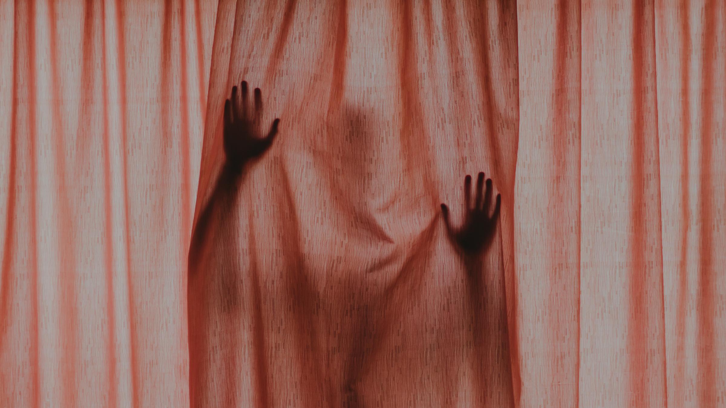 Eine Gestalt mit erhobenen Händen hinter einem verschlossenen roten Vorhang