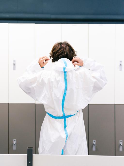 Ein Mensch in Krankenhausschutzbekleidung von hinten vor Schließfächern, die Kapuze über den Kopf ziehend.