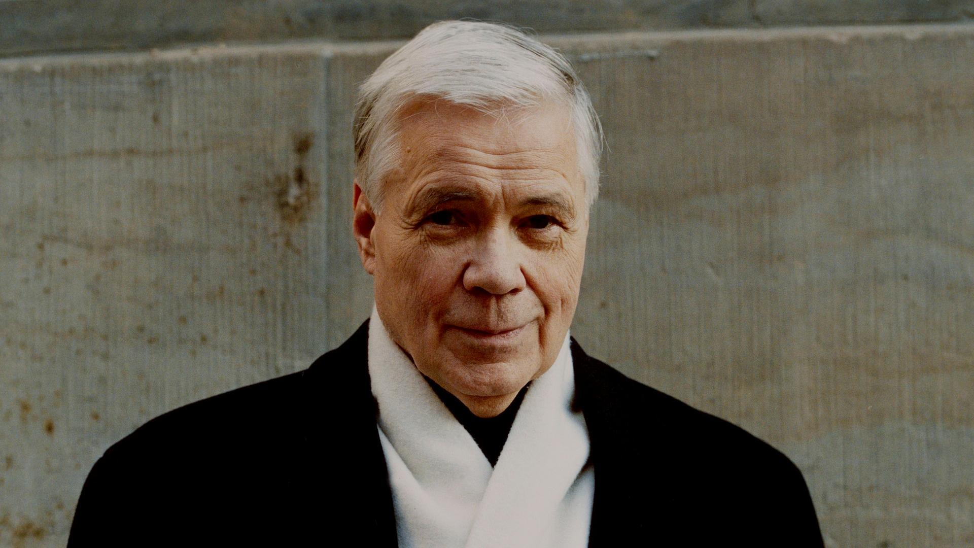 Dietrich Fischer-Dieskau steht in dunklem Mantel und mit weißem Schal vor einer Wand. Er hat weiße Haare und lächelt verschmitzt in die Kamera.