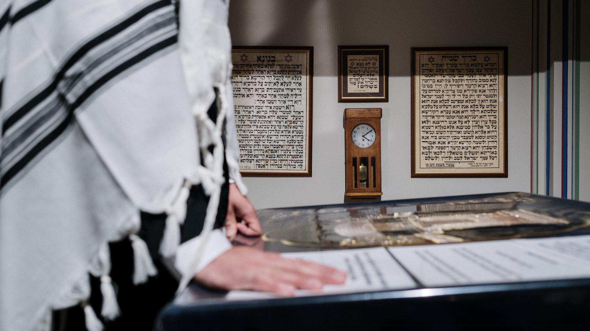 Eine Person mit einem über die Schultern geworfenen Tallit steht vor einer hebräischen Schrift. Eine Person mit einem über die Schultern geworfenen Tallit steht vor einer hebräischen Schrift.