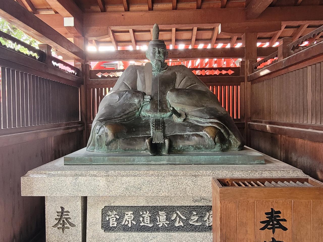 Die Bronze-Statue des Gelehrten Sugawara no Michizane steht unter einem Holzdach, durch das Licht fällt, auf einem Steinsockel.