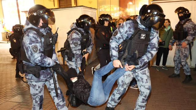 Drei Polizisten mit Helmen tragen einen Mann in schwarzer Kleidung an Händen und Füßen weg.