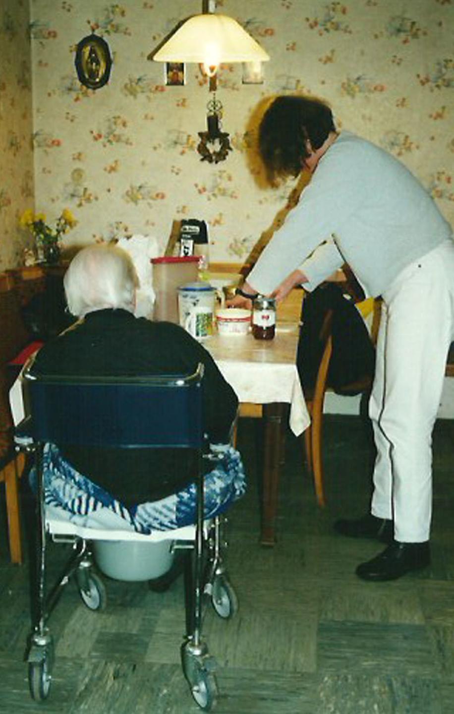 Eine ältere Frau sitzt in einem Rollstuhl an einem Esstisch. Eine Krankenschwester bereitet das Essen vor. Eine ältere Frau sitzt in einem Rollstuhl an einem Esstisch. Eine Krankenschwester bereitet das Essen vor.
