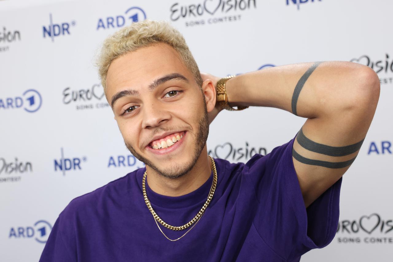 Malik Harris lächelt in die Kamera. Er trägt ein lila T-Shirt. Eine Goldkettte schmückt seinen Hals. Malik Harris lächelt in die Kamera. Er trägt ein lila T-Shirt. Eine Goldkettte schmückt seinen Hals.