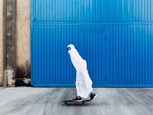 Ein Kind in einem Gespensterkostüm mit blauer Maske um die Augen fährt auf einem Skateboard an einem blauen Tor vorbei.