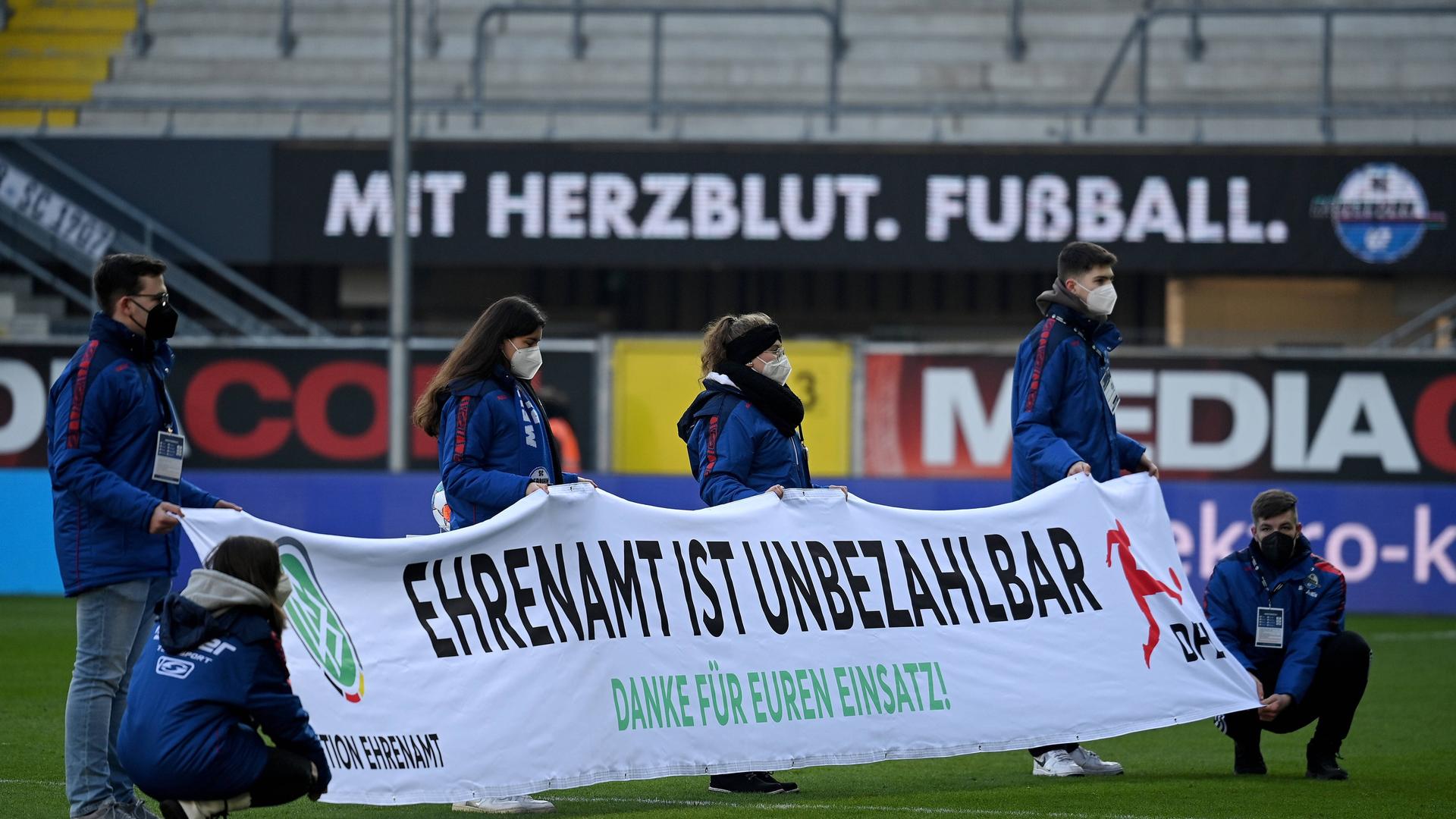 Freiwillige halten in einem Fußballstadion ein Banner hoch mit der Aufschrift: Ehrenamt ist unbezahlbar.