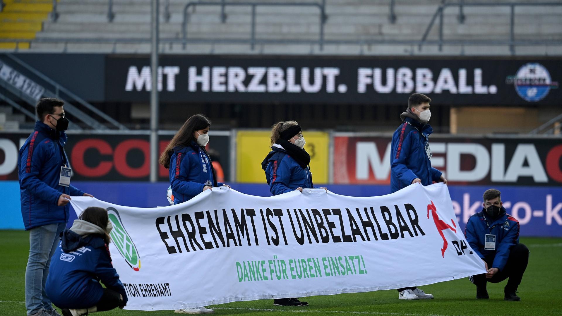 Freiwillige halten in einem Fußballstadion ein Banner hoch mit der Aufschrift: Ehrenamt ist unbezahlbar. Freiwillige halten in einem Fußballstadion ein Banner hoch mit der Aufschrift: Ehrenamt ist unbezahlbar.