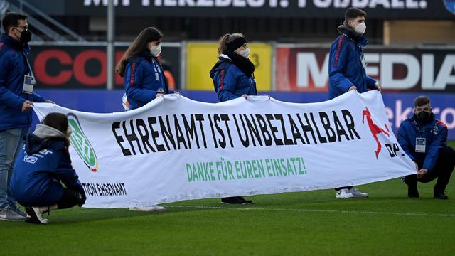 Freiwillige halten in einem Fußballstadion ein Banner hoch mit der Aufschrift: Ehrenamt ist unbezahlbar.