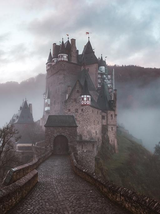 Graf Dracula entführt Prof. van Dusen in sein Schloss. Wird der Professor sich auch diesmal befreien können? Zu sehen: Ein Weg zu einer Burg mit Türmen. Im Hintergrund Gebirge mit Nebel und dichten Wolken. 
