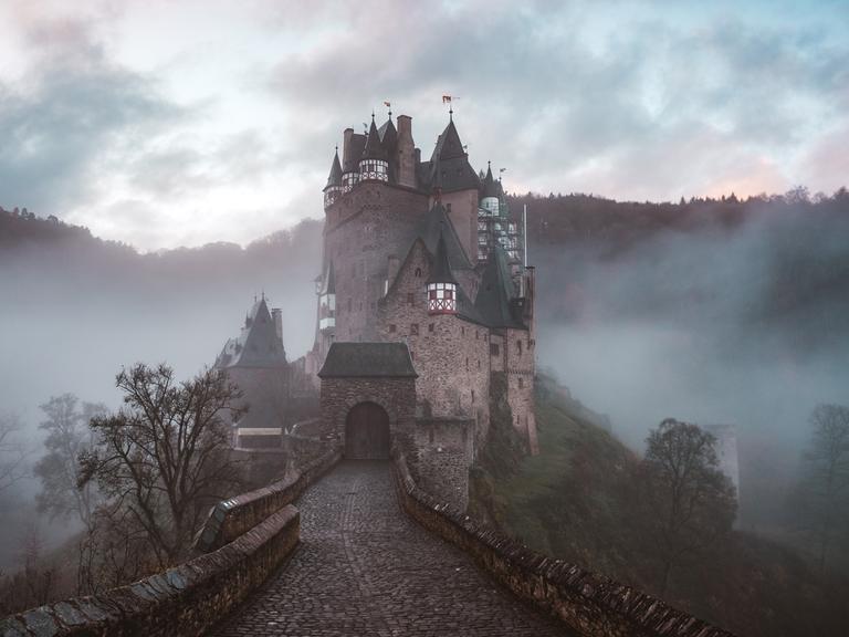 Graf Dracula entführt Prof. van Dusen in sein Schloss. Wird der Professor sich auch diesmal befreien können? Zu sehen: Ein Weg zu einer Burg mit Türmen. Im Hintergrund Gebirge mit Nebel und dichten Wolken. 