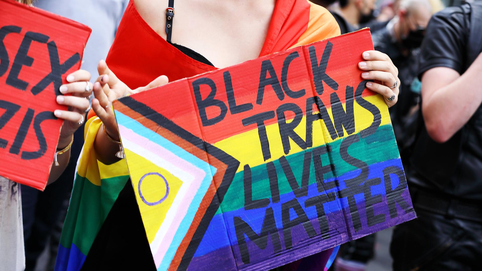 Demoteilnehmerin mit umgehangener Regenbogenfahne trägt Protestschild mit der Aufschrift: Black Trans Lives Matter Demoteilnehmerin mit umgehangener Regenbogenfahne trägt Protestschild mit der Aufschrift: Black Trans Lives Matter