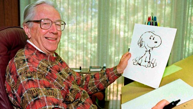 Charles Schulz hält eine Zeichnung von Snoopy in der Hand und lächelt.