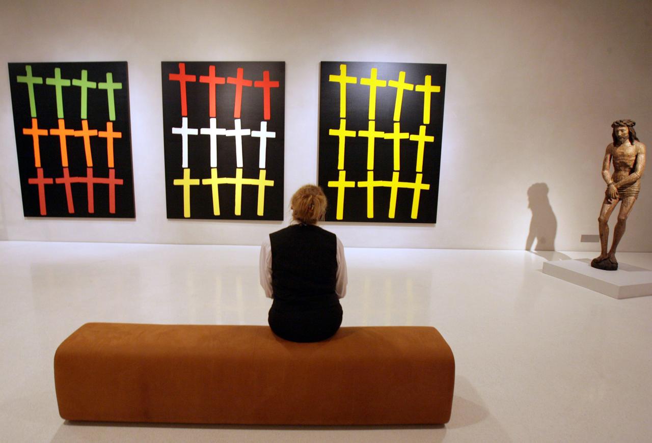 Ein Museumsbesucher blickt auf Bilder von Andy Warhol, die Kreuze zeigen. Rechts eine Christusfigur.  