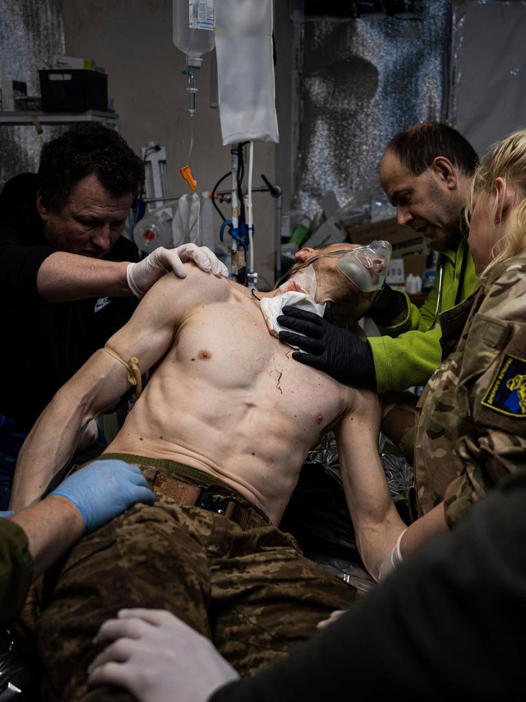 Ein verwundeter Mann ist umgeben von Ärzten und Pflegern, die ihn versorgen. Er trägt eine Camouflage-Hose, ebenso wie eine Frau, die rechts von ihm steht mit dem Rücken zur Kamera. Der Oberkörper des Mannes ist frei.