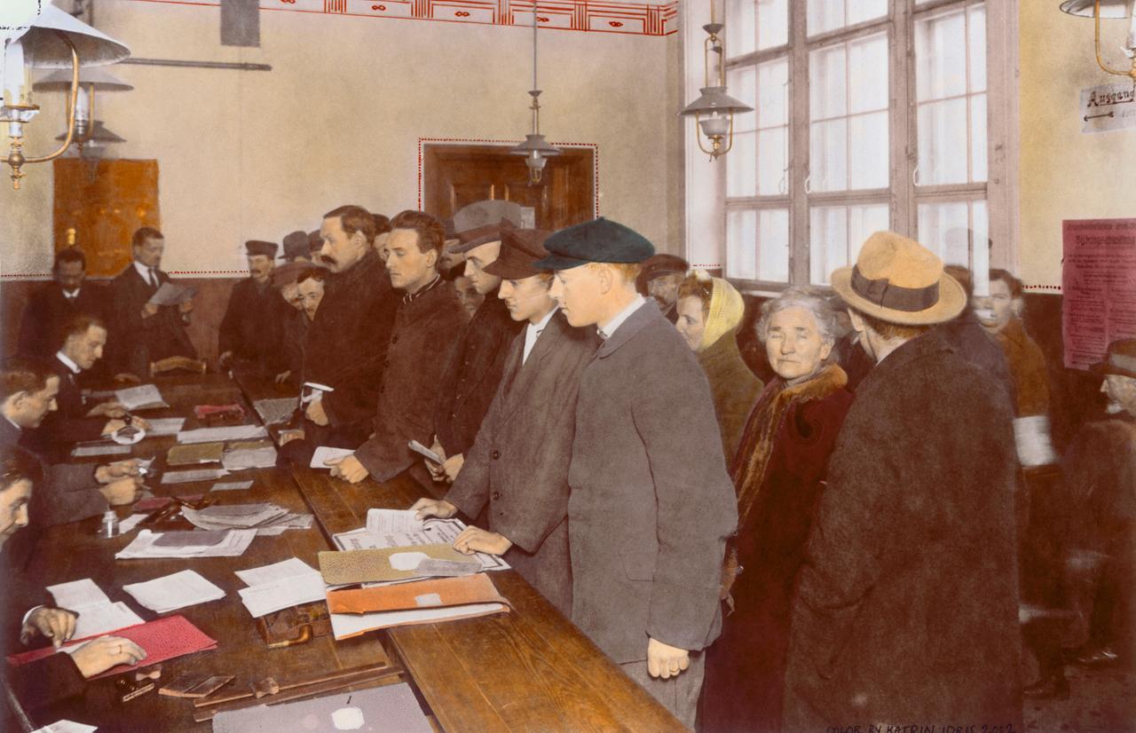 Mehrere Personen stehen Schlange vor einem Tisch an dem ihnen die Erwerbslosenfürsorge ausgezahlt wird. Das Bild wurde nachträglich koloriert.