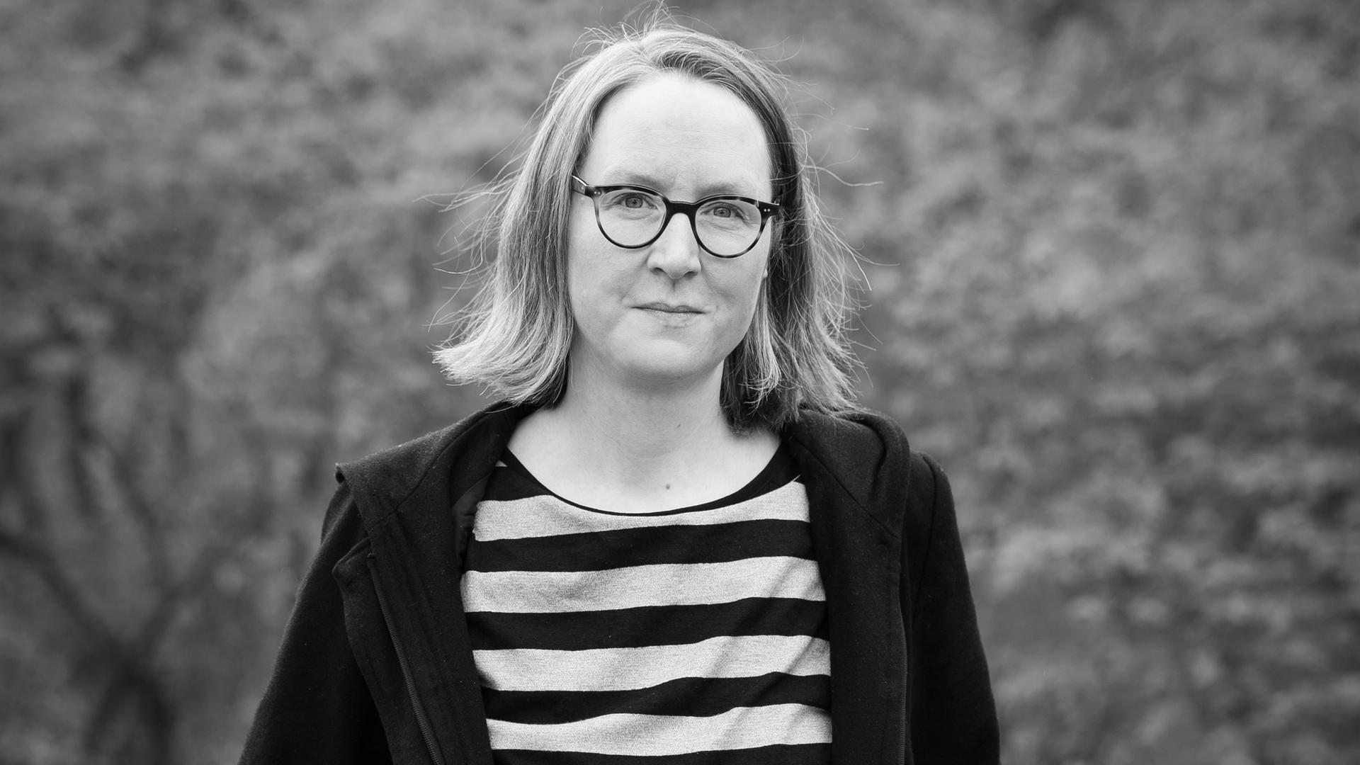 Friederike Schmitz schaut aud dem Schwarz-Weiß-Foto in die Kamera. Sie hat schulterlange Haare und trägt Brille. Sie hat ein quergestreiftes Oberteil an und darüber eine dunkle Jacke. Friederike Schmitz schaut aud dem Schwarz-Weiß-Foto in die Kamera. Sie hat schulterlange Haare und trägt Brille. Sie hat ein quergestreiftes Oberteil an und darüber eine dunkle Jacke.