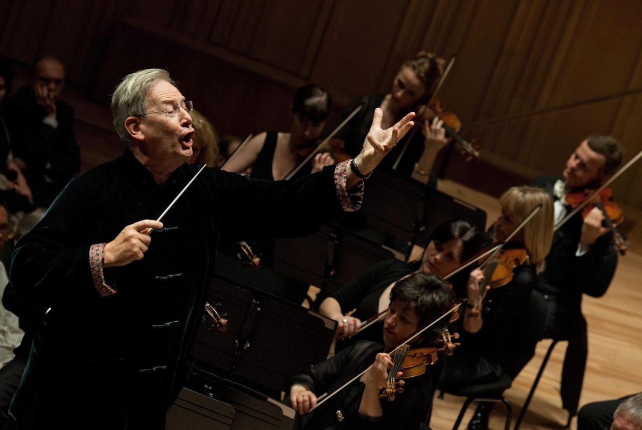 Der Dirigent Sir John Gardiner steht mit auffordernder Geste vor einem Orchester und scheint mitzusingen, um dem Chor den richtigen Einsatz zu geben. Der Dirigent Sir John Gardiner steht mit auffordernder Geste vor einem Orchester und scheint mitzusingen, um dem Chor den richtigen Einsatz zu geben.